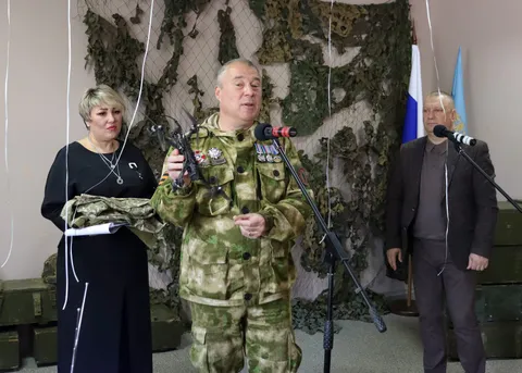Гость в военной форме выступает у микрофона, ведущая держит переданную музею форму